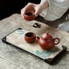 Person holding a teacup from a Yixing Clay Gongfu Tea Set, with a Yixing Gongfu Tea Set teapot and another cup on a bamboo-patterned tea tray on a rustic wooden table