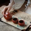 Hands adjusting the teapot of a Yixing Clay Gongfu Tea Set, with two teacups from the Yixing Gongfu Tea Set placed on a bamboo-patterned tea tray atop a rustic wooden table, showcasing traditional gongfu tea preparation