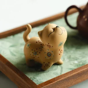 Yixing Cat tea pet ornament, a handmade golden clay cat tea pet with colorful floral patterns, displayed on a fabric-covered tea tray with a Yixing teapot in the background for a traditional gongfu tea setup