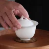 Hand touching the lid of a 100 ml white porcelain gaiwan, placed on a wooden tray with a natural fiber coaster