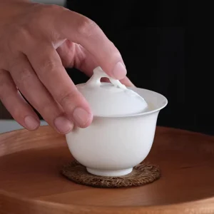 Hand touching the lid of a 100 ml white porcelain gaiwan, placed on a wooden tray with a natural fiber coaster