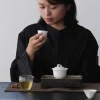 Person sipping tea from a small cup, with a 100 ml white porcelain gaiwan on a dark tea tray, alongside a glass pitcher and teacups
