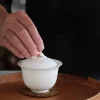 Close-up of a hand touching the lid of a 100 ml white porcelain gaiwan on a wooden tray
