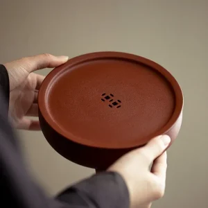 Home 29 Hands holding a round Yixing Zhuni water storage tea tray, showing the smooth surface and hollow drainage design of the zisha gongfu tea tray