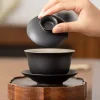 Lifting the lid of a Black Gaiwan to reveal its smooth interior, placed on a wooden stand to showcase its minimalist craftsmanship.
