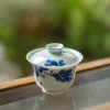 Dehua Gaiwan with blue floral motifs, placed on a glass surface with a greenery background for a serene display.