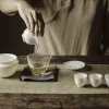 Pouring steeped tea from a white porcelain vessel into a glass fairness pitcher, part of a Gaiwan Porcelain tea set.