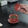 Lifting the lid of a red Yixing Clay Gaiwan to reveal steeping tea leaves, capturing the ritual of traditional gongfu tea brewing.