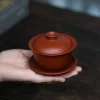 Holding a bright red Gaiwan Handmade from Yixing clay, emphasizing its ergonomic design and artisanal craftsmanship for daily use.