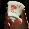 Holding a teacup from the Red Blossom Gaiwan collection, revealing the maker’s mark on its base while showcasing the vibrant plum blossom artwork