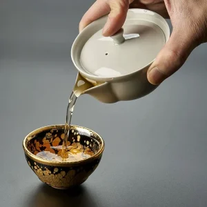 Pouring tea from a Gaiwan With Spout into a decorative gold-spotted teacup, demonstrating its functional and elegant design.
