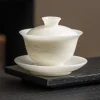 Mutton Fat Jade Gaiwan placed on a dark tea tray, its milky white porcelain and relief landscape details creating a serene, minimalist tea setup with the gaiwan tea bowl.