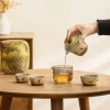 Pouring tea from the Travel Gaiwan into a glass pitcher, with three teacups ready, showcasing the gongfu tea brewing process with this portable travel tea set.