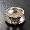 Complete Titanium Gaiwan with lid resting on a lotus leaf saucer, featuring a sleek metallic finish and ergonomic wooden knob.