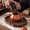 A hand pouring loose tea leaves into a Yixing Pumpkin Teapot, which rests on a carved wooden tea tray.